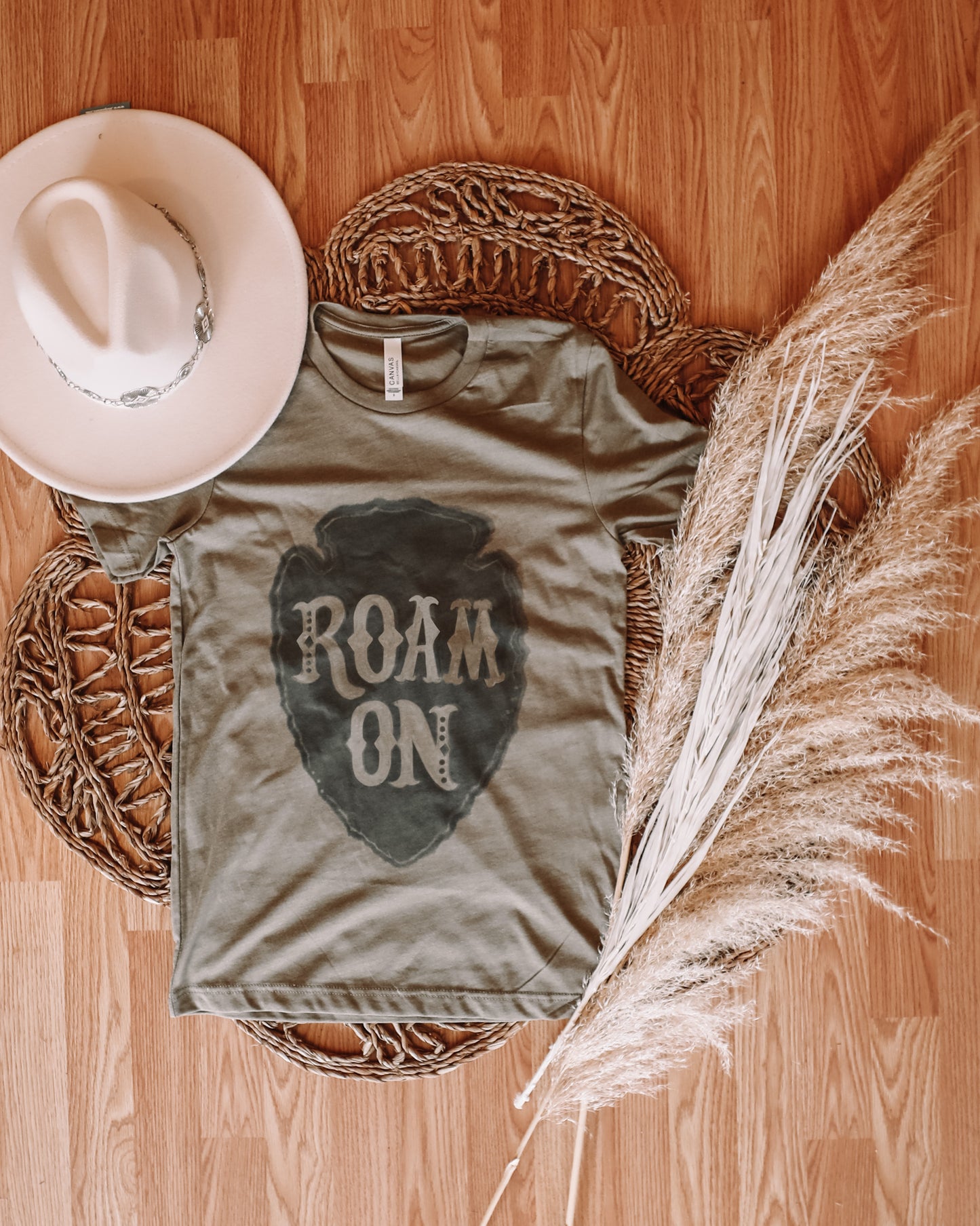 Roam On Western graphic tee in army green featuring an arrowhead design photographed in a flatlay form with pampas grass rug and western concho hat